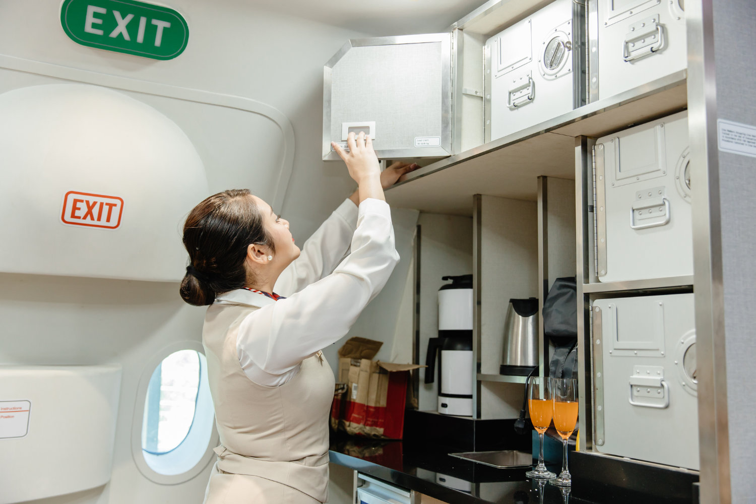 Flight attendant accessing a storage cabinet in an aircraft cabin, with visible safety exit signs and refreshments on the counter.