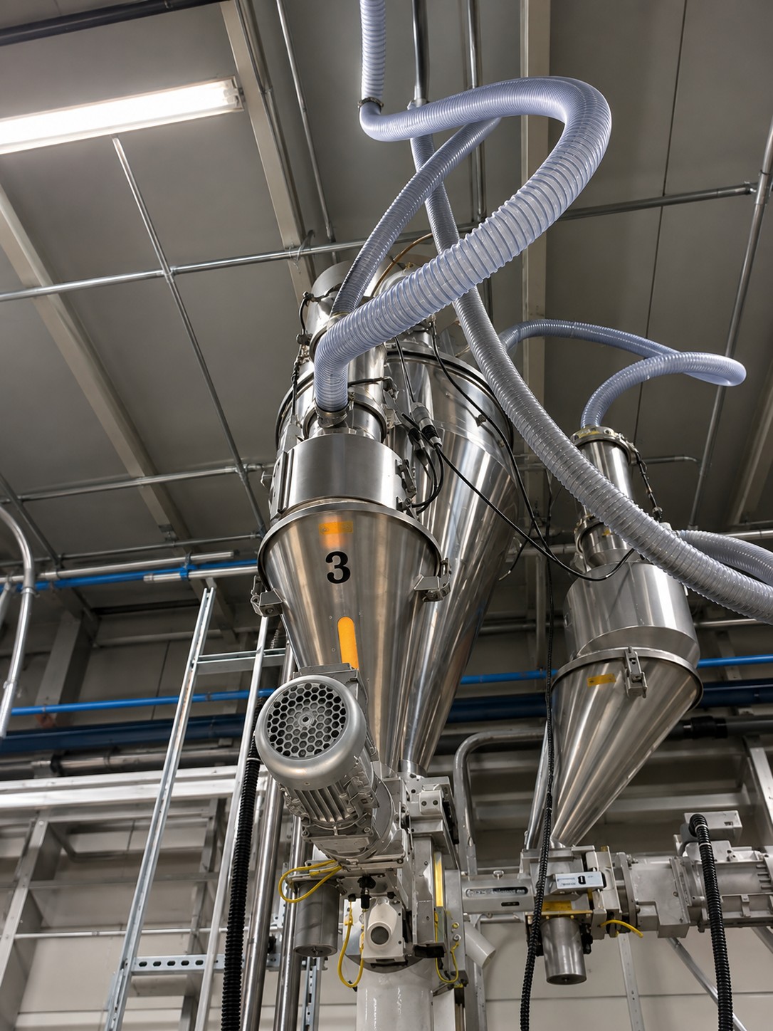 Twin screw extrusion machinery with interconnected metal hoppers and flexible tubing in an industrial setting.
