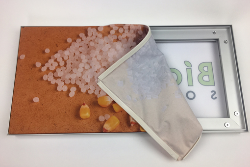Plastic pellets and corn kernels on a surface, partially covered by a fabric displaying the word "Bio" from BioPlastic Solutions.